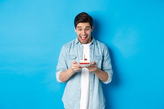 Excited young male model looking at birthday cake and smiling, making a wish and celebrating, standing against blue background