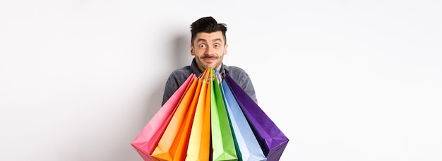 Возбужденный молодой человек носит красочные сумки для покупок и улыбается счастливому покупателю, покупающему на распродаже, стоя на