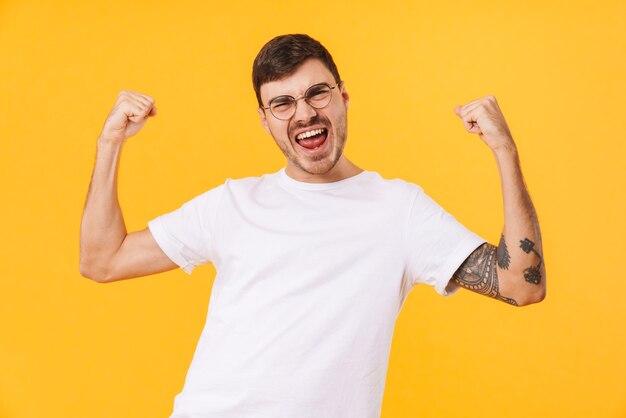 excited young man in eyeglasses making winner gesture isolated over yellow wall