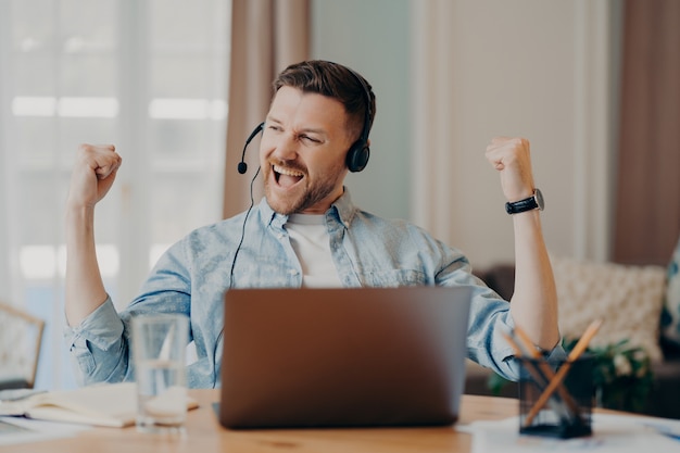 Eccitato giovane auricolare freelancer con microfono che celebra il successo dopo un'importante riunione online o colloquio di lavoro, seduto alla scrivania nel soggiorno di casa. concetto di comportamento vincente