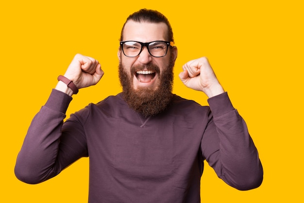 Excited young man is screaming because is excited with both hands with glasses near a yellow wall