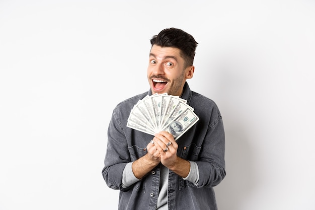 Excited young man smiling and showing dollar bills, making money, standing against white background.