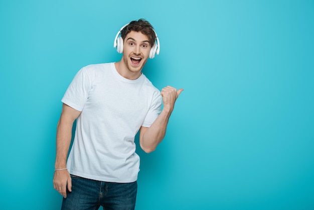 Excited young man in wireless heaphones pointing with thumb while looking at camera on blue