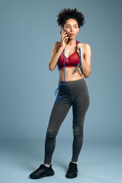 Excited young multiracial woman wearing sportswear talking at the smartphone after fitness while holding jumping rope at her neck