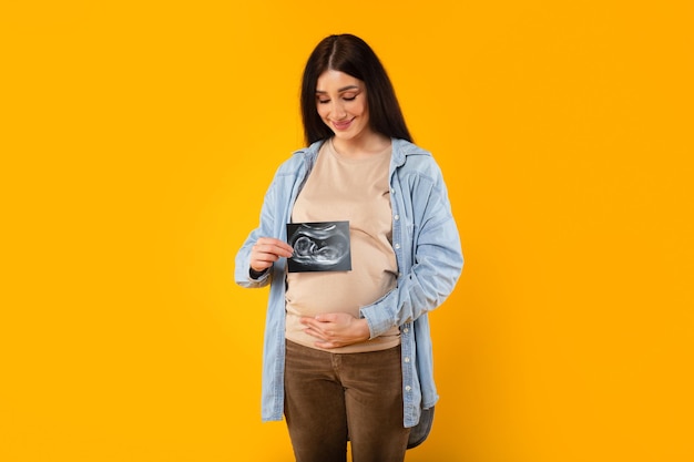 Excited young pregnant woman embracing belly and showing ultrasound picture of unborn baby on yellow