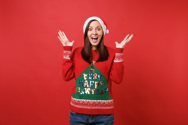 Eccitato giovane ragazza santa in cappello di natale che tiene la bocca spalancata, guardando le mani spalancate sorprese isolate su sfondo rosso. felice anno nuovo 2019 celebrazione festa concetto. mock up copia spazio.