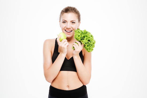 Excited young sports woman holding lettuce and green apple isolated    