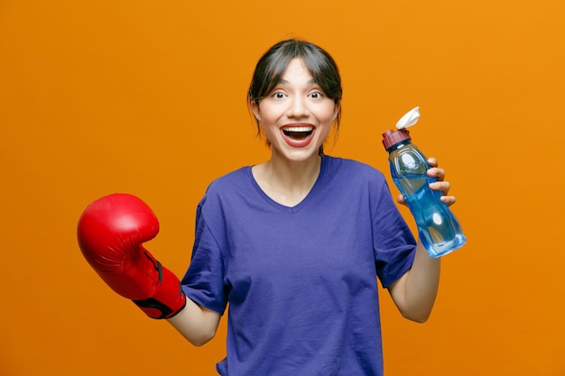 excited young sporty woman wearing tshirt and boxing gloves looking at camera holding water bottle keeping hand in air isolated on orange background