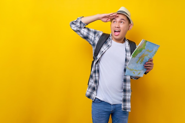 Excited young traveler tourist asian man in casual clothes hat\
holds city map during the excursion looking aside on yellow\
background passenger traveling on weekend air flight journey\
concept