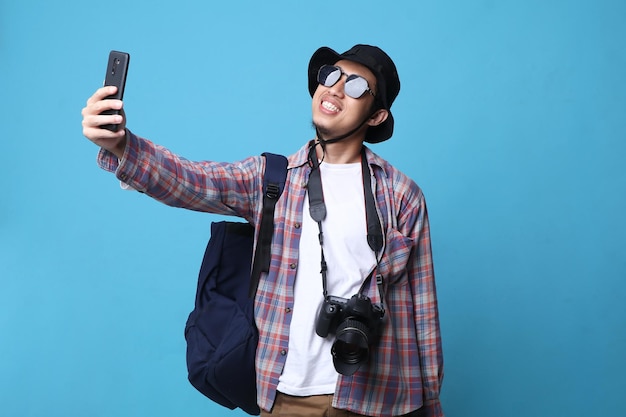 青の背景に携帯電話で自撮りをしている帽子をかぶった興奮した若い旅行者の観光客の男性