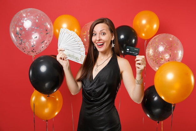 Excited young woman in black dress celebrating holding credit card, bundle lots of dollars, cash money on red background air balloons. Women's Day Happy New Year birthday mockup holiday party concept.