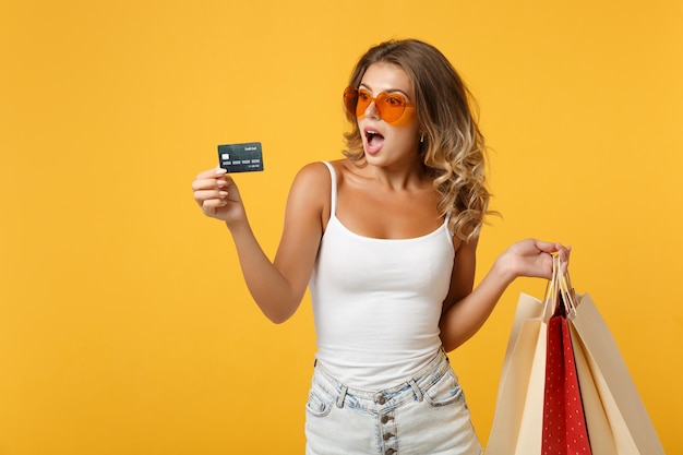 Ragazza eccitata della giovane donna in occhiali che posano isolata su fondo giallo arancio. concetto di stile di vita delle persone. mock up spazio di copia. tenendo la borsa del pacchetto con gli acquisti dopo lo shopping, carta di credito.