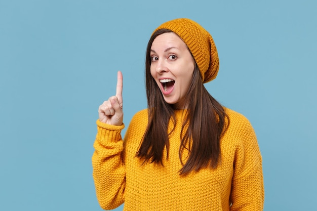 Excited young woman girl in yellow sweater and hat posing\
isolated on blue background studio portrait. people emotions\
lifestyle concept. mock up copy space. hold index finger up with\
great new idea.