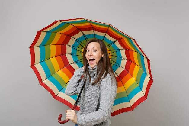 Excited young woman in gray sweater, scarf keeping mouth open holding colorful umbrella isolated on grey background. Healthy fashion lifestyle people emotions, cold season concept. Mock up copy space.