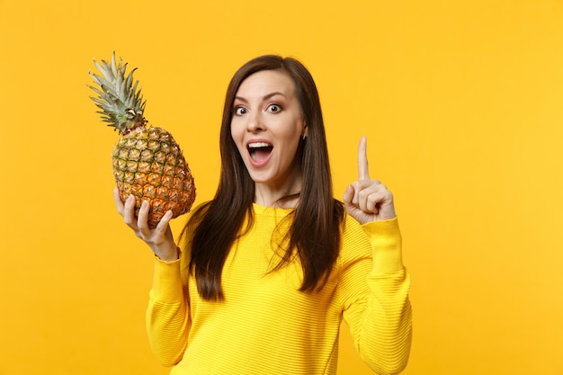 Giovane donna eccitata che tiene la bocca aperta, puntando il dito indice verso l'alto con una nuova grande idea che tiene frutta fresca di ananas isolata su sfondo giallo arancio. persone stile di vita vivido, relax concetto di vacanza