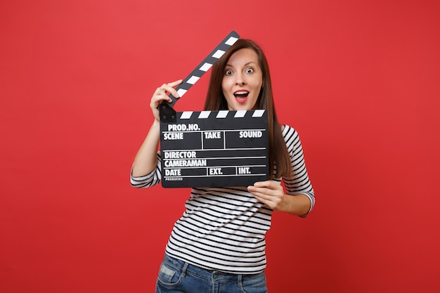 Excited young woman keeping mouth wide open looking surprised holding classic black film making clapperboard isolated on red background. People sincere emotions, lifestyle concept. Mock up copy space.