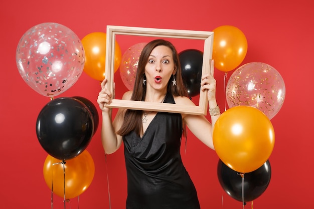 Eccitata giovane donna in abitino nero che celebra e tiene in mano una cornice su mongolfiere di sfondo rosso brillante. giornata internazionale della donna, felice anno nuovo, concetto di festa di compleanno mockup.