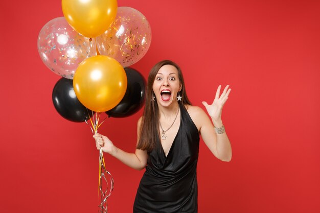 Возбужденная молодая женщина в маленьком черном платье празднует разведение рук, держа воздушные шары, изолированные на красном фоне. Международный женский день, с новым годом, концепция праздничной вечеринки макет дня рождения.