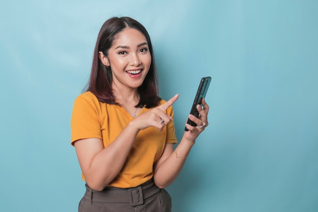 An excited young woman pointing her smartphone in her hand isolated on blue background