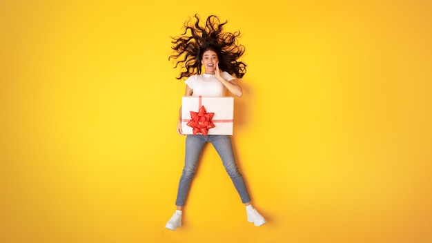 Excited young woman posing with wrapped gift box yellow background