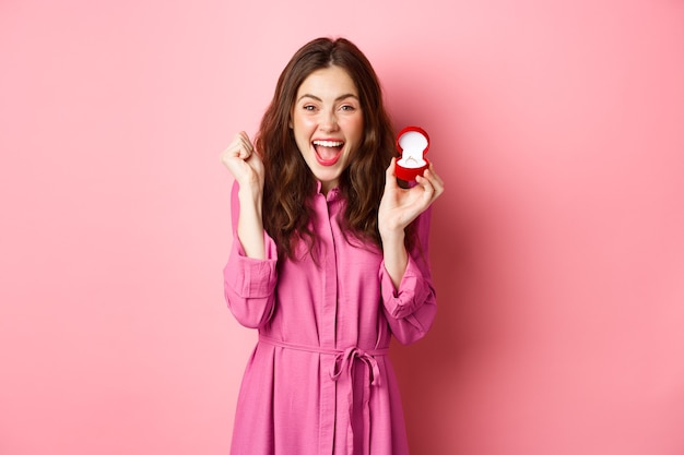 Excited young woman showing engagement ring and celebrating getting married, smiling happy, talking about wedding day, receive marriage proposal, standing over pink wall. Copy space