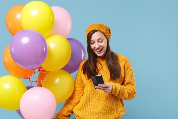 青い背景に分離されたポーズのセーター帽子で興奮した若い女性。誕生日の休日のパーティー、人々の感情の概念。コピースペースをモックアップします。携帯電話を使ってカラフルな風船を持ってお祝いしましょう。