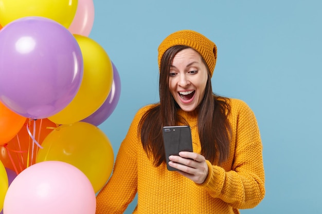 Excited young woman in sweater hat posing isolated on blue\
background. birthday holiday party, people emotions concept. mock\
up copy space. celebrating hold colorful air balloons using mobile\
phone.