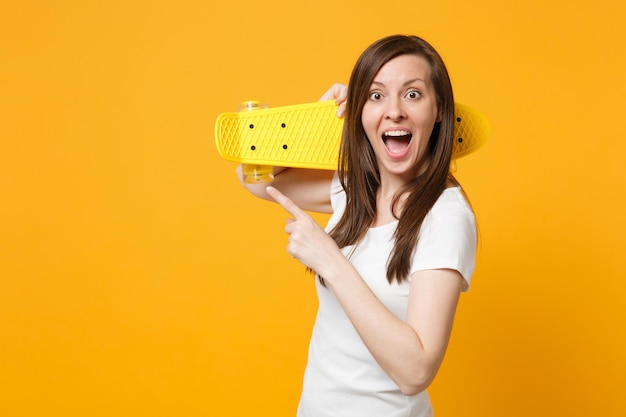 Excited young woman in white casual clothes pointing index finger aside, holding yellow skateboard isolated on bright yellow orange background in studio. People lifestyle concept. Mock up copy space.