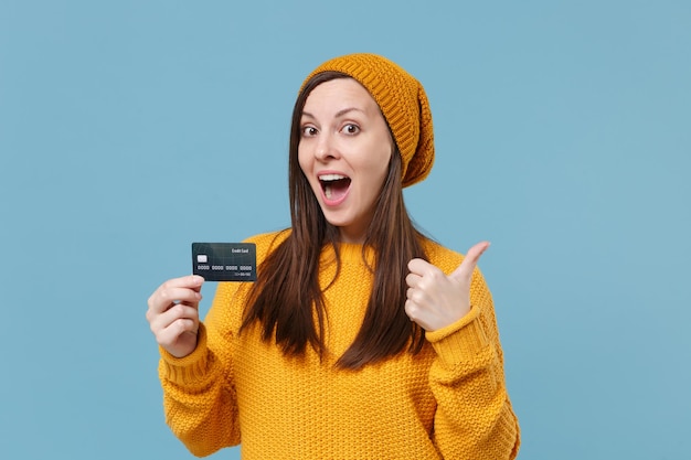 Excited young woman in yellow sweater and hat posing isolated\
on blue background studio portrait. people sincere emotions\
lifestyle concept. mock up copy space. showing thumb up hold credit\
bank card.