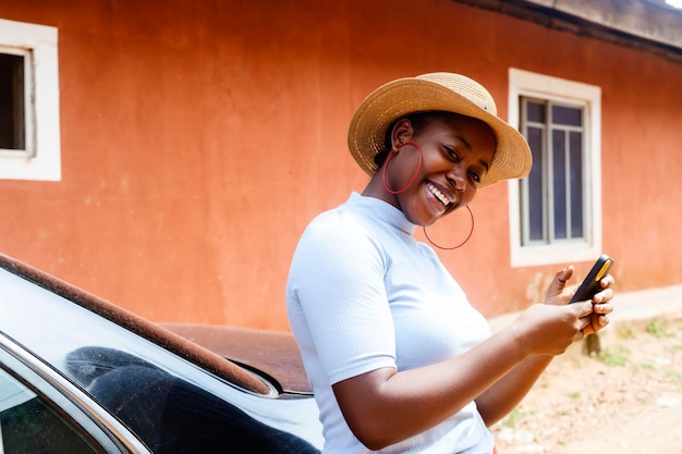 Exciting black african female entrepreneur holding smart phone in hand while standing near modern car outdoors