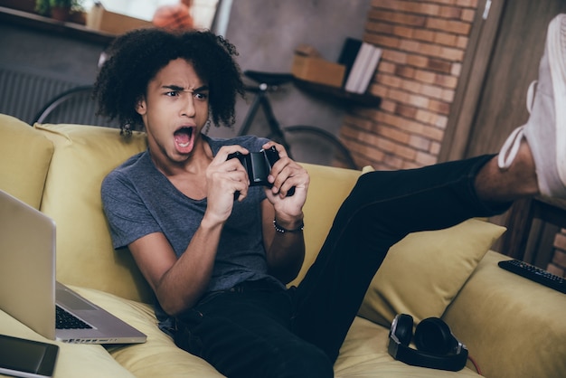 Photo exciting game! excited young african man playing video game and keeping his mouth open while sitting on the couch at home