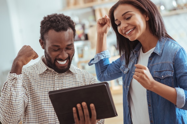 Exciting news. Joyful young man and woman standing in the cafe and raising hands in celebration, having read an email with good news on tablet