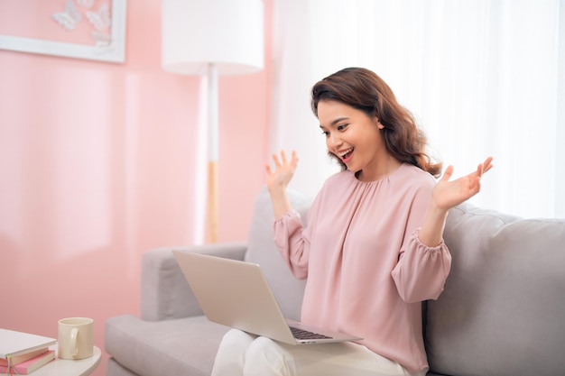 Exciting young Asian woman using laptop when sitting on sofa at home