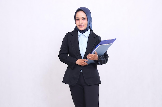 An exclusive Asian office woman wearing a hijab stands cheerfully carrying a pen clipboard and noteb