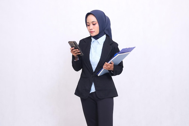 Exclusive Asian office woman wearing a hijab stands elegantly operating a smartphone and carrying a