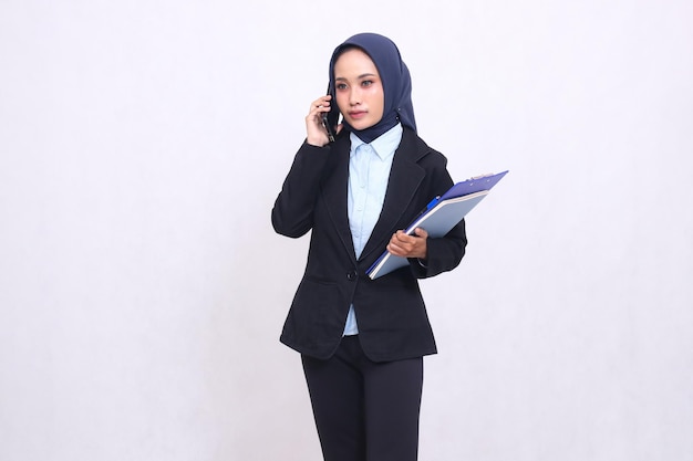 Exclusive Asian office woman wearing a hijab stands elegantly on the phone and carries a pen clipboa