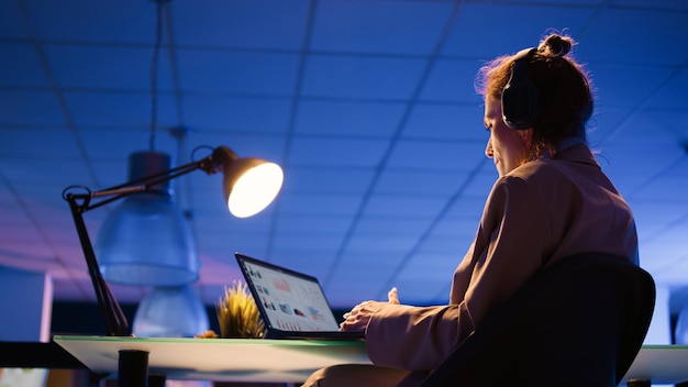 Executive assistant listening to music late at night, wearing headset and enjoying work. Young employee with heeadphones analyzing financial graphs report with analytics. Handheld shot.