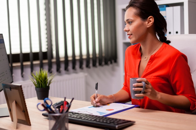 Executive businesswoman entrepreneur writing financial sales report looking at screen wearing red. Successful financial auditor in data marteking looking at graphs on computer screen and writing repor