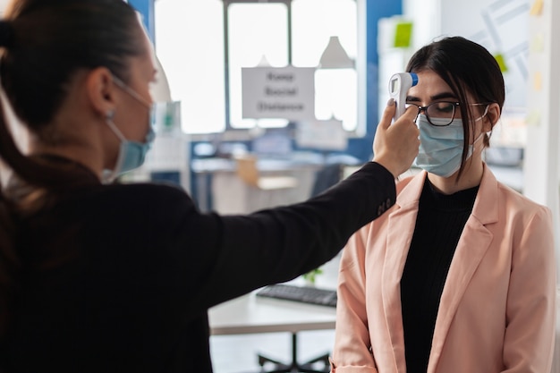 Photo executive manager measuring temperature using infrared medical thermometer to prevent infection with coronavirus before entering in startup company office. businesswoman with protection face mask