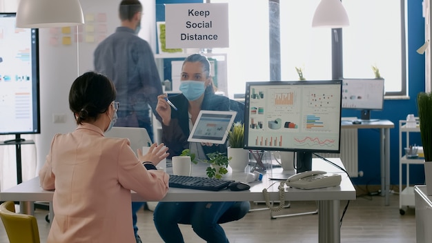 Executive manager with face mask showing statistics to her collegue using digital tablet computer. Coworkers working in new normal office keeping ing ing social distance to prevent infection with covi