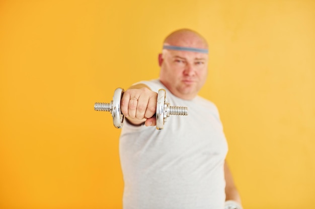 Photo exercises by using dumbbells funny overweight man in sportive head tie is against yellow background