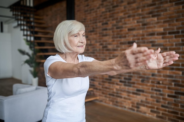 Exercising. Gray-haired senior woman standing with her arms forward