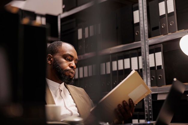 Exhausted african american detective solving complex crime case and reading folder with police reports. Tierd investigator working late, studying archival records information