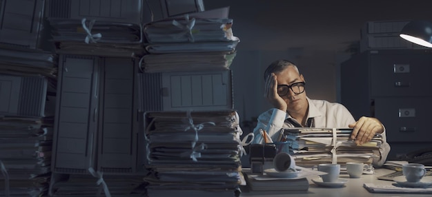 机で寝て夜遅くまで残業して疲れ果てたビジネスマンが書類の山に囲まれている