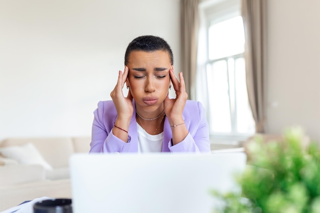 ホームオフィスで頭痛を抱えている疲れ果てた女性オフィスの机で働くアフリカ系アメリカ人の創造的な女性が疲れを感じているストレスのたまったビジネスウーマンが過労中に目の痛みを感じている