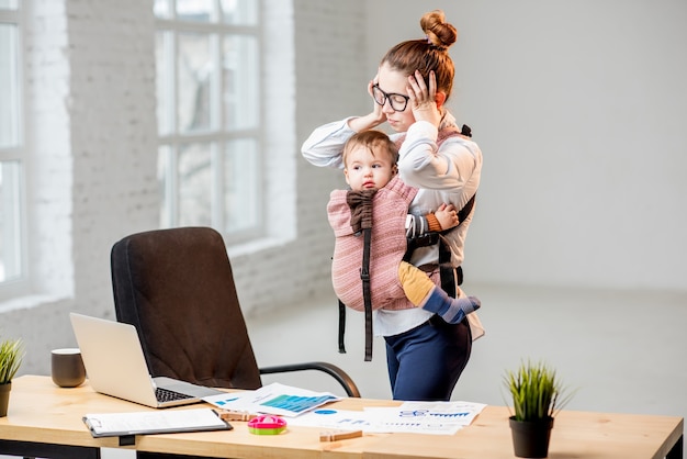 Photo exhausted businesswoman holding her head standing with her baby son at the office during the work