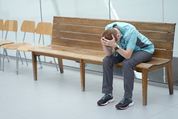 Exhausted caucasian female doctor is upset due to patient loss after a long shift, sitting on a banch in a hospital corridor and hoding herself head