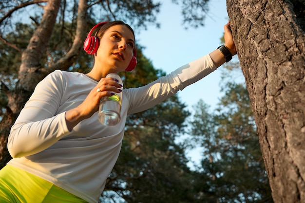 Photo exhausted female runner does not feel well due to dehydration or overtraining young lady drinks water from a sports bottle after a hard workout leaning on a tree trunk