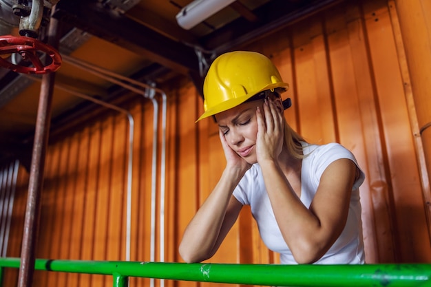 Exhausted female worker with protective helmet on head leaning on railing in factory and having headache because she is overworked.