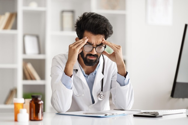 Photo exhausted hindu man doctor sitting at desk touching head